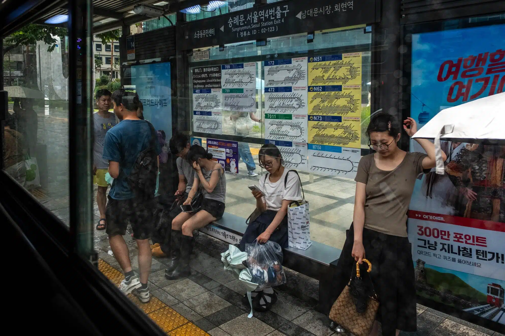 seoul-streets-july-2026-23 People wait at a bus stop in central Seoul under heavy monsoon rain in July 2026.